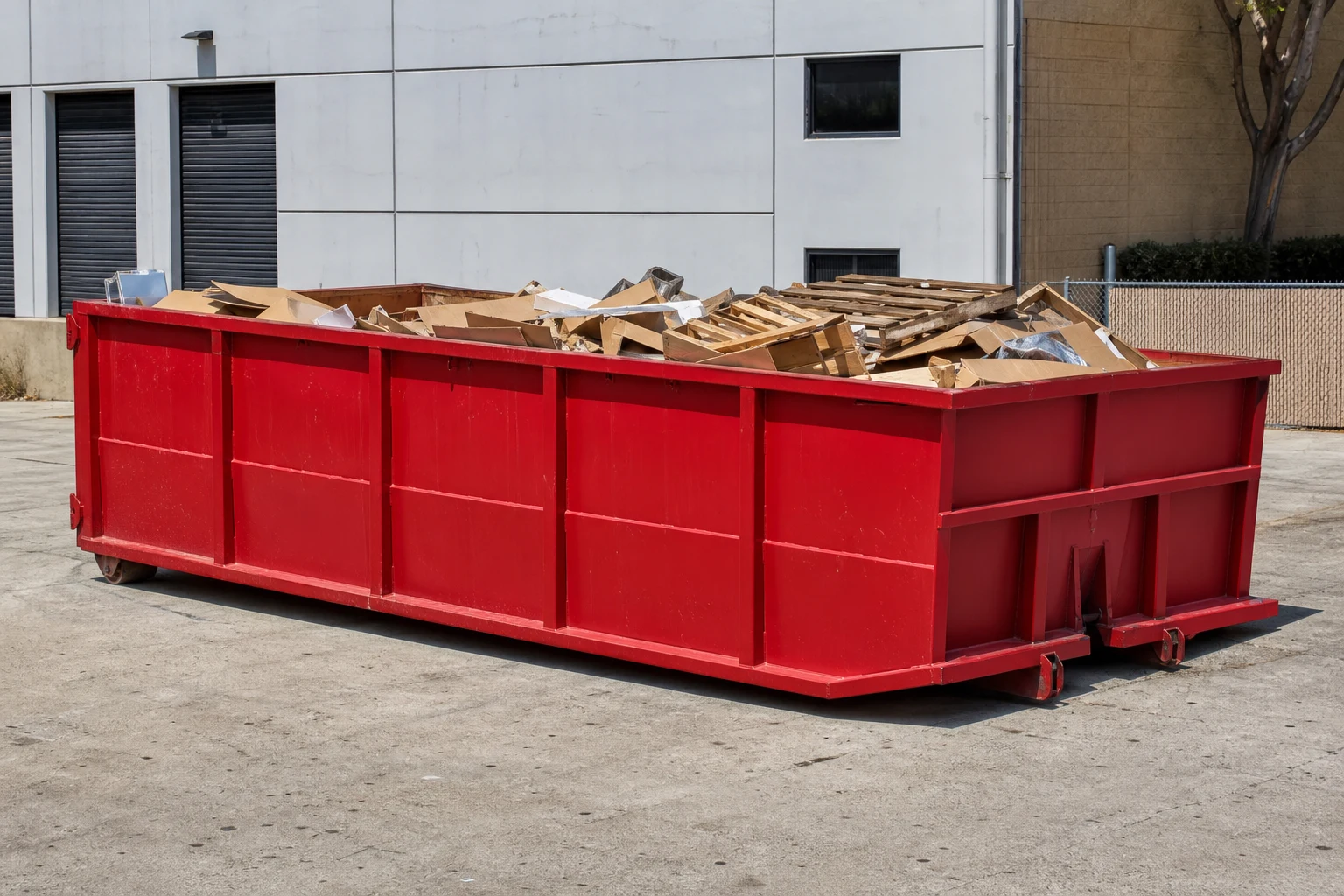 Red roll-off dumpster beside staged cardboard pallets and business cleanout debris near a light commercial building